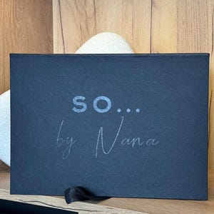 May include: A dark blue rectangular box with the text "so... by Nana" in a light grey font. The box has a black ribbon tied to the bottom right corner. The box is on a wooden surface.