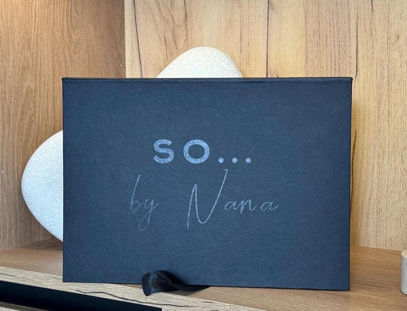 May include: A dark blue rectangular box with the text "so... by Nana" in a light grey font. The box has a black ribbon tied to the bottom right corner. The box is on a wooden surface.