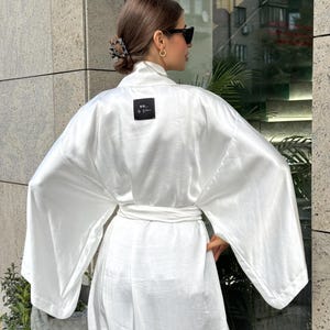 May include: A white satin robe with wide sleeves and a matching belt. The robe features a small black square label with white text that reads "SO... by Nina". The robe is worn outdoors.