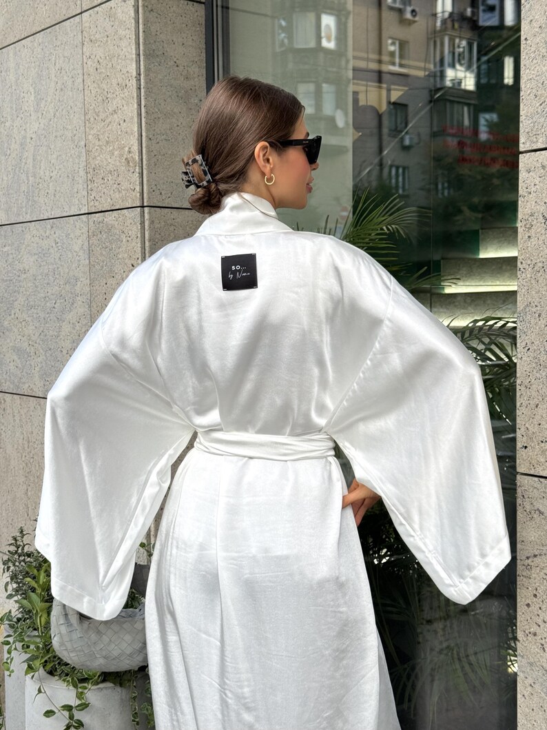 May include: A white satin robe with wide sleeves and a matching belt. The robe features a small black square label with white text that reads "SO... by Nina". The robe is worn outdoors.
