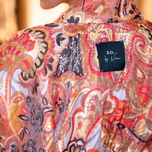 May include: A patterned garment in red, orange, and brown, featuring a paisley-style design with floral and abstract shapes. A black label with the text "so... by Nana" is sewn on the back of the garment.