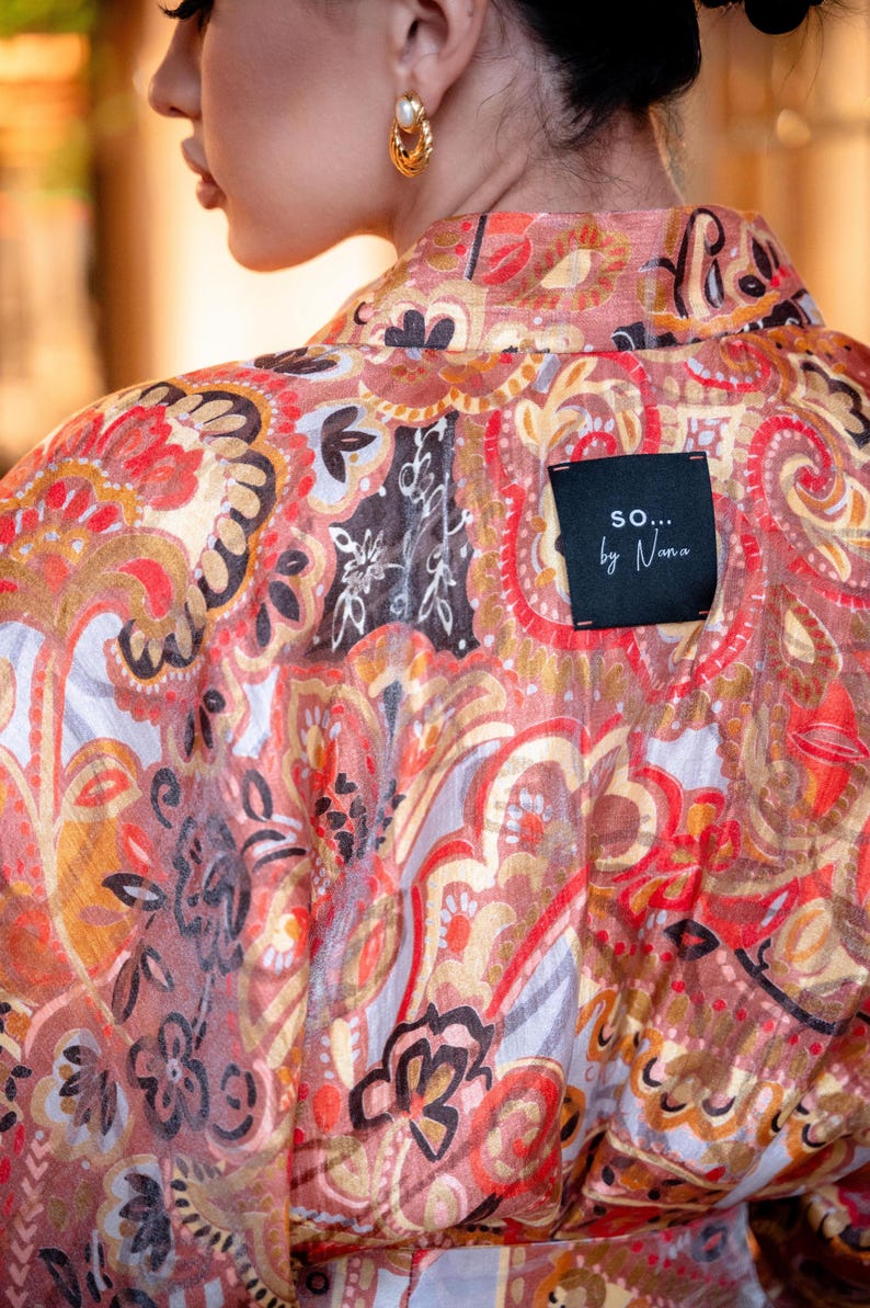 May include: A patterned garment in red, orange, and brown, featuring a paisley-style design with floral and abstract shapes. A black label with the text "so... by Nana" is sewn on the back of the garment.