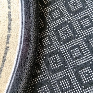 May include: A close-up view of the underside of a rug featuring a black non-slip backing with a repeating white square pattern. The edge of the rug is finished with a thick, black stitched border that separates the backing from the beige top surface.