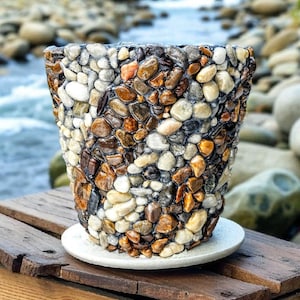May include: A decorative planter made of small, colorful river stones. The stones are various shades of brown, gray, and white, creating a textured surface. The planter sits on a white saucer, placed on a wooden surface.