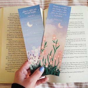 May include: Two floral bookmarks with inspirational quotes are placed in an open book. One bookmark reads "God is within her, she will not fall" and the other says "Charm is deceitful and beauty is vain, but a woman who fears the Lord is to be praised."
