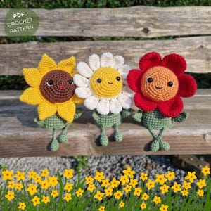 Op de afbeelding: Drie gehaakte bloemen amigurumi speelgoedjes die op een houten bankje zitten. De bloemen zijn een gele zonnebloem, een witte madeliefje en een rode klaproos. Elke bloem heeft een glimlachend gezicht en groene benen.