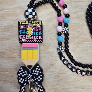 May include: A lanyard featuring black beads, colorful silicone accents, and a square charm with the word TEACHER repeated in pink, blue, and yellow with a lightning bolt. It includes a pencil-shaped charm, a black and white checkered bow, and beads spelling MRS SMI with a heart.