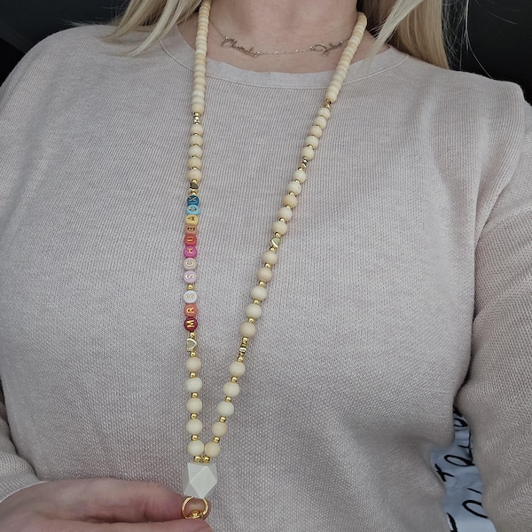 May include: A lanyard featuring light-colored wooden beads interspersed with small gold-toned metallic beads. One side includes a vertical sequence of colorful letter beads spelling out MRS SCHUBACK, finished with a large white geometric bead.