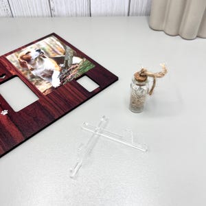 May include: A wooden photo frame with a picture of a beagle dog, two empty photo slots, and paw print details. A small glass bottle with a cork stopper and twine is next to the frame. Two clear plastic stands are in front.