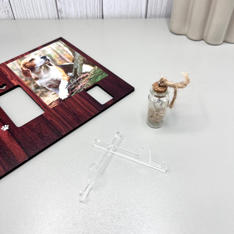 May include: A wooden photo frame with a picture of a beagle dog, two empty photo slots, and paw print details. A small glass bottle with a cork stopper and twine is next to the frame. Two clear plastic stands are in front.