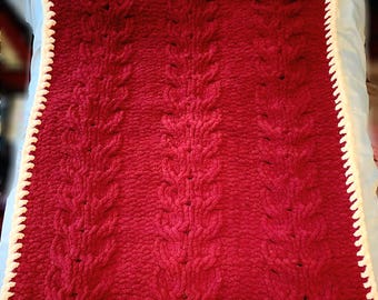 May include: A handmade, red knitted blanket with a cable knit design. The blanket has a white, scalloped border. The texture of the blanket is soft and plush, perfect for snuggling. The blanket is a cozy home decor item.
