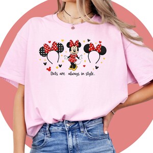 May include: A light pink t-shirt featuring a Minnie Mouse design. The design includes Minnie Mouse, two sets of ears with bows, and the text "Dots are always in style." The Minnie Mouse character is wearing a red dress with white polka dots.