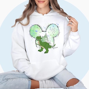 May include: White hoodie with a green dinosaur inside a Mickey Mouse ear outline. The dinosaur is standing in front of a castle with fireworks in the background. The word "Rex" is written in cursive below the dinosaur.