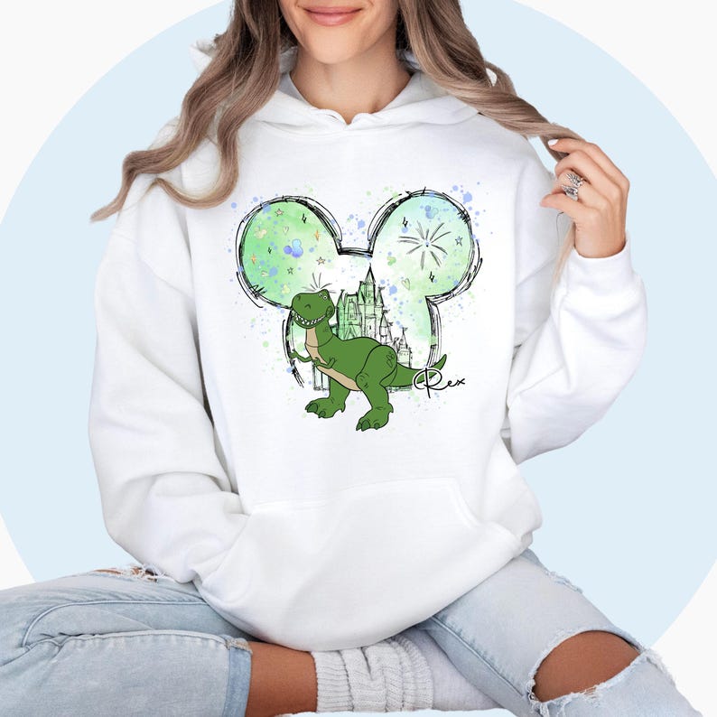 May include: White hoodie with a green dinosaur inside a Mickey Mouse ear outline. The dinosaur is standing in front of a castle with fireworks in the background. The word "Rex" is written in cursive below the dinosaur.
