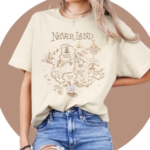 May include: A beige t-shirt featuring a brown graphic of a Neverland map. The map includes landmarks such as Skull Rock, Mermaid Lagoon, and Pirate Cove. The text "Neverland" is printed in brown above the map.