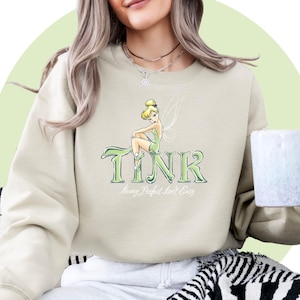 May include: A light beige sweatshirt with a graphic print of Tinkerbell sitting on the word "TINK" with the text "Being Perfect Isn't Easy" underneath.
