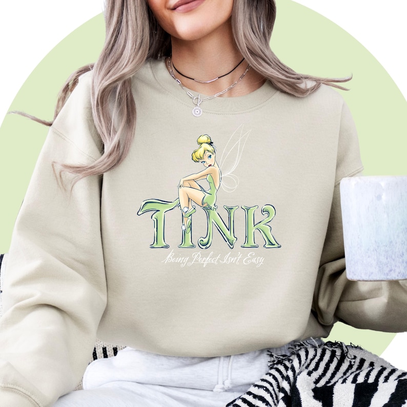 May include: A light beige sweatshirt with a graphic print of Tinkerbell sitting on the word "TINK" with the text "Being Perfect Isn't Easy" underneath.