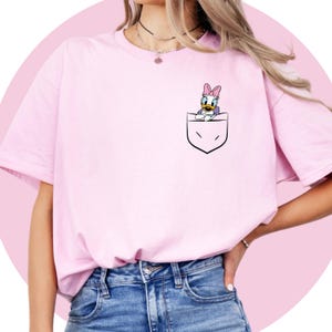 May include: A pink t-shirt with a cartoon duck character in a pocket on the front. The duck is wearing a pink bow.