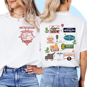 May include: White t-shirt with a red and white graphic of Lightning McQueen from the movie Cars. The back of the shirt features a map of Radiator Springs with various landmarks and businesses, including Luigi's Casa Della Tires, Cozy Cone Motel, and Fillmore's.