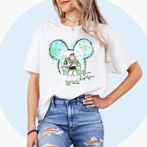 May include: White t-shirt with a green, blue, and white watercolor Mickey Mouse design featuring Buzz Lightyear from Toy Story. The text "Buzz Lightyear" is written in black below the design.