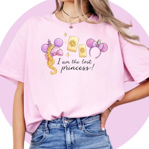May include: A pale pink t-shirt with a princess-themed graphic. The design includes a tower, a braid, lanterns, and a headband with mouse ears. The text "I am the lost princess!" is printed on the shirt.