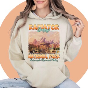 May include: A beige hoodie with a vintage-style graphic of Radiator Springs National Park, a fictional town from the Disney Pixar movie Cars. The graphic features the text "Radiator Springs" and "National Park" with the tagline "Gateway to Monument Valley".