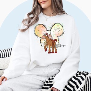 May include: White sweatshirt with a watercolor Mickey Mouse silhouette featuring a cartoon horse from the Toy Story movie. The horse is brown with a red saddle and the text "Bullseye" is written below the horse.