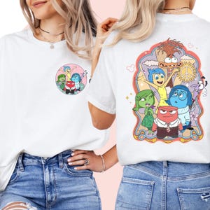 Könnte beinhalten: Zwei weiße T-Shirts mit einem Grafikdruck der Figuren aus dem Film Inside Out. Die Grafik auf der Vorderseite des Shirts zeigt die Figuren in einem Kreis, während die Grafik auf der Rückseite des Shirts die Figuren in einem aufwendigeren Design mit einem Peace-Zeichen und einer Wolke zeigt.