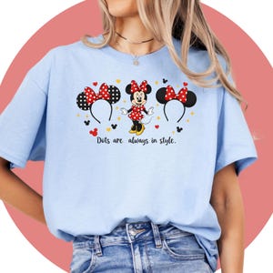 May include: Light blue t-shirt featuring a Minnie Mouse graphic. The design includes Minnie Mouse, polka dot bows, and the text "Dots are always in style." The shirt is made of a soft material.