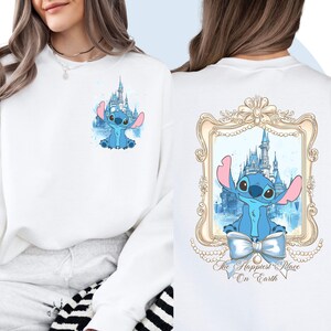 Puede incluir: Sudadera blanca con un personaje azul Stitch y un castillo en el fondo. La parte posterior de la sudadera tiene un marco dorado con un personaje azul Stitch y un castillo en el fondo. El texto "The Happiest Place On Earth" est&aacute; debajo del marco.