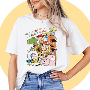May include: White t-shirt with a vibrant cartoon graphic of characters from a well-known animated film. The phrase "YOU'VE GOT A FRIEND IN ME" is printed above the characters in a playful font. The design includes a dinosaur, a cowboy, and a space ranger.