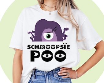 Puede incluir: Camiseta blanca con un diseño gráfico que presenta una criatura morada con un gran ojo verde y el texto "SCHMOOPSIE POO" en negro. El diseño está centrado en la parte delantera de la camiseta.