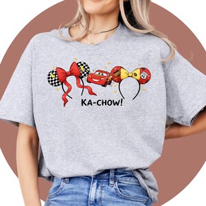 May include: A light gray t-shirt featuring a design of cartoon cars and mouse ears. The design includes a red bow, a red car, and the text "KA-CHOW!". The mouse ears are black and white checkered. The shirt is made of a soft material.