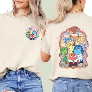 Op de afbeelding: Een licht beige T-shirt met een grafische print van personages uit de film Inside Out. De personages zijn Joy, Sadness, Anger, Disgust en Fear. De grafiek bevindt zich op de achterkant van het shirt. De voorkant van het shirt heeft een kleinere grafiek van de personages.
