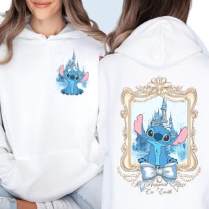 Puede incluir: Sudadera blanca con un personaje azul Stitch delante de una imagen de acuarela del castillo de Cenicienta. La parte posterior de la sudadera tiene un marco dorado con una imagen de acuarela del castillo de Cenicienta y Stitch delante. El texto "The Happiest Place On Earth" est&aacute; debajo del marco.