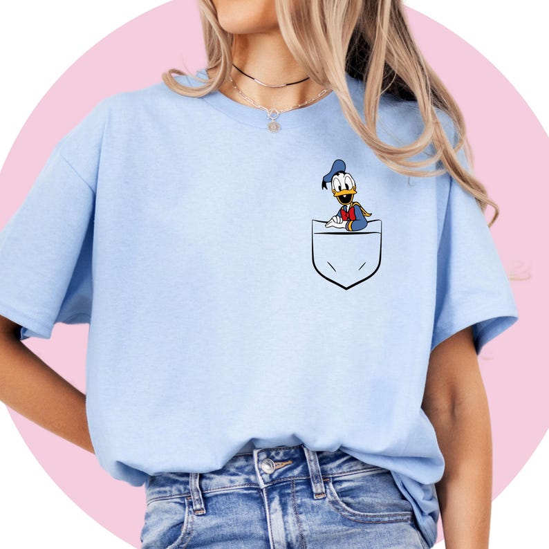May include: A light blue t-shirt with a cartoon image of Donald Duck peeking out of a pocket on the front of the shirt.