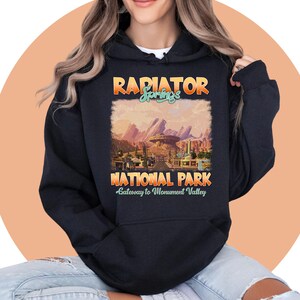 May include: Black hooded sweatshirt with a vintage-style graphic design featuring the text "Radiator Springs National Park" and a scenic image of a desert landscape with a red rock formation. The text "Gateway to Monument Valley" is also included.