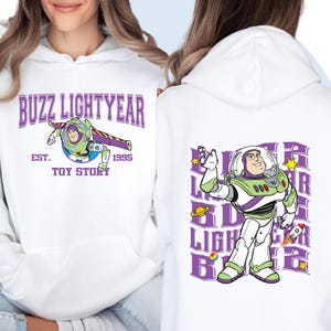May include: White hoodie featuring Buzz Lightyear graphics from Toy Story. The front displays "BUZZ LIGHTYEAR" with an image of Buzz in his space suit, the text "EST. 1995 TOY STORY". The back shows a larger Buzz Lightyear graphic.