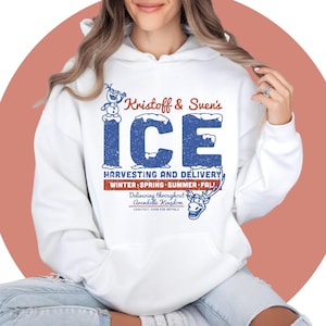Pode incluir: Moletom branco com um gráfico azul e vermelho. O design diz "Kristoff & Sven's ICE Harvesting and Delivery" com um boneco de neve, rena e as estações "Winter, Spring, Summer, Fall."