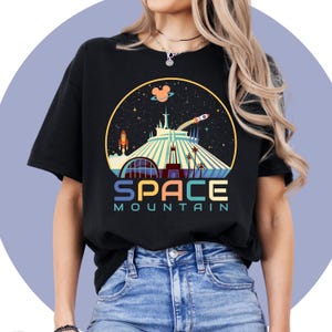 以下が含まれることがあります： ディズニーワールドのスペースマウンテンのカラフルなグラフィックがプリントされた黒いTシャツ。グラフィックにはロケット、ミッキーマウス、星が含まれています。グラフィックの下には「SPACE MOUNTAIN」というテキストがプリントされています。