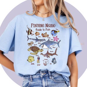 May include: Light blue t-shirt featuring a graphic from the movie "Finding Nemo." The design includes various cartoon fish characters with their names, such as Nemo, Dory, and Bruce. The text "Finding Nemo Guide to Fish" is printed at the top.