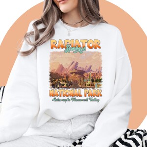 May include: White sweatshirt with a vintage-style graphic of Radiator Springs National Park from the movie Cars. The graphic features a sunset scene with mountains and buildings. The text "Radiator Springs National Park" and "Gateway to Monument Valley" is printed in a retro font.