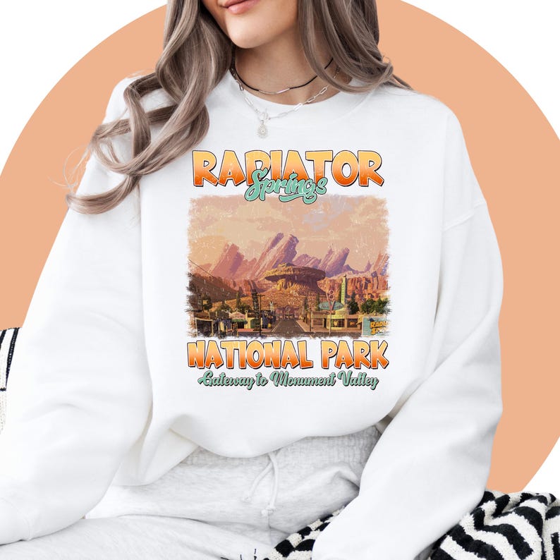 May include: White sweatshirt with a vintage-style graphic of Radiator Springs National Park from the movie Cars. The graphic features a sunset scene with mountains and buildings. The text "Radiator Springs National Park" and "Gateway to Monument Valley" is printed in a retro font.