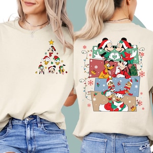 May include: A beige t-shirt with a Christmas tree design featuring Mickey Mouse, Minnie Mouse, Donald Duck, Goofy, and Pluto. The tree is decorated with red, green, and yellow ornaments. The back of the shirt features a cartoon design of Mickey Mouse, Minnie Mouse, Goofy, and Donald Duck.