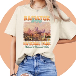May include: A beige t-shirt with a vintage-style graphic of the "Radiator Springs National Park" with the text "Gateway to Monument Valley".