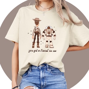 Puede incluir: Una camiseta beige con una imagen de dibujos animados de Woody y Buzz Lightyear de Toy Story. La camiseta tiene el texto "You got a friend in me" impreso.