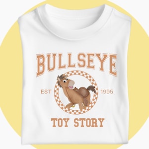 May include: White t-shirt with a cartoon image of Bullseye from Toy Story. The design includes the word "BULLSEYE" in a brown, collegiate-style font above a circular graphic of the horse. The text "EST 1995" and "TOY STORY" are also included.