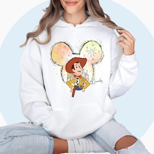 May include: White hoodie with a colorful Mickey Mouse silhouette featuring a cartoon image of Woody from Toy Story. The silhouette includes a fireworks display and the word "Woody" is written in cursive.