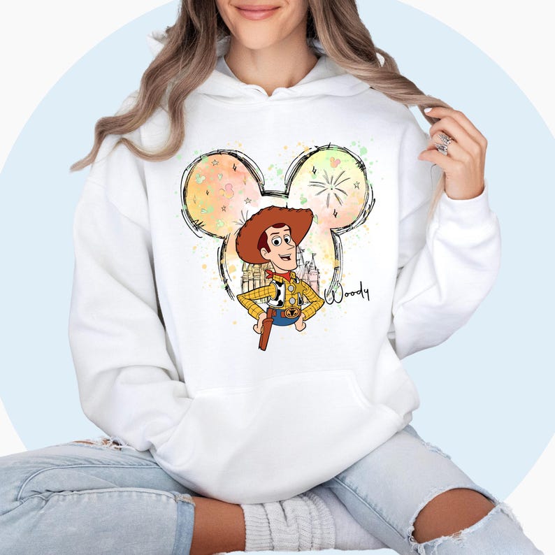 May include: White hoodie with a colorful Mickey Mouse silhouette featuring a cartoon image of Woody from Toy Story. The silhouette includes a fireworks display and the word "Woody" is written in cursive.