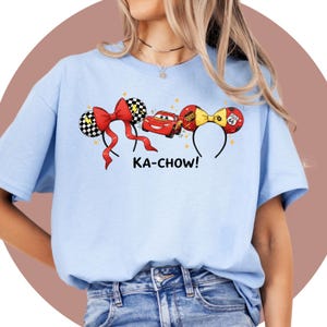 May include: Light blue t-shirt featuring a graphic of cartoon race cars and themed headbands. The design includes a red bow, checkered ears, and the text "KA-CHOW!". The shirt is made of a soft material.